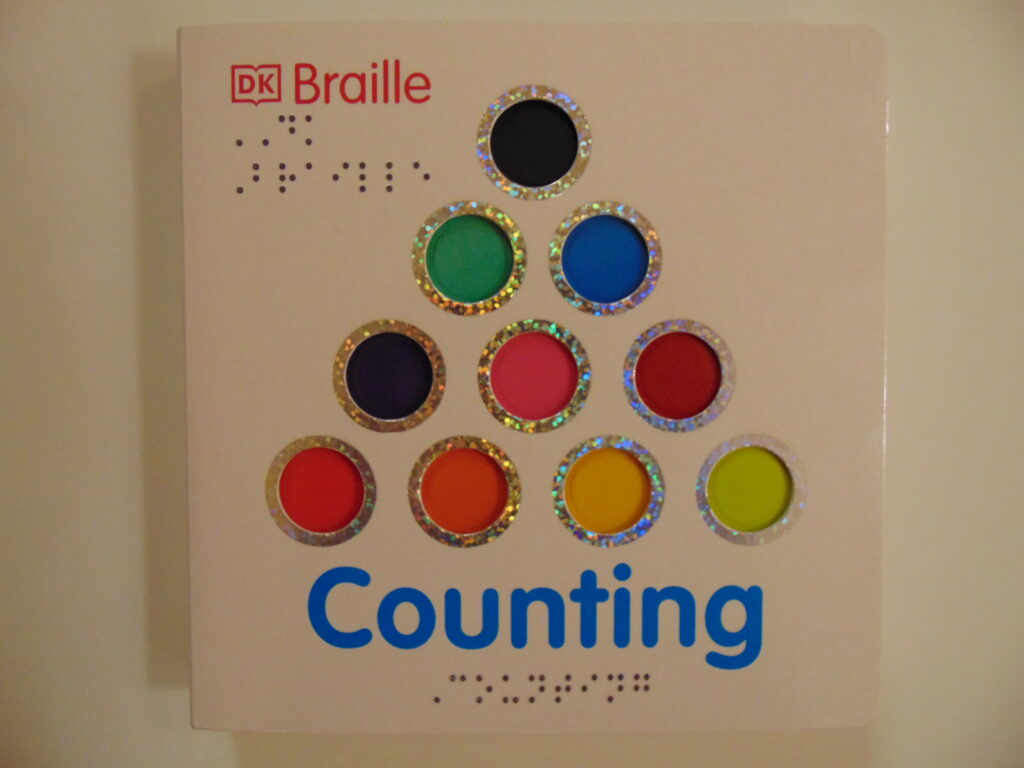 Image description:  Front cover with a white background, “DK” with an outline of a book around it and “Braille” in red print and the braille translation underneath in the top left corner, In the middle is a pyramid shape with ten dots of a variety of colors with sparkly silver outlines, underneath in blue print and braille is the word “Counting”